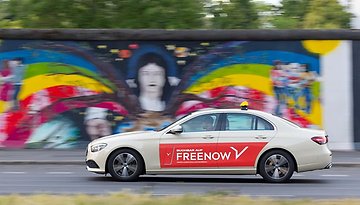 A beige car with a red 'FREENOW' logo drives past a colorful mural.