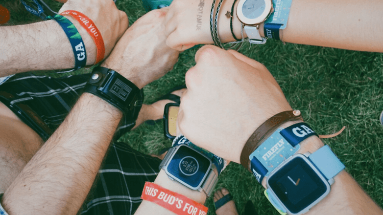 A group of hands wearing various colorful wristbands and smartwatches on grass.