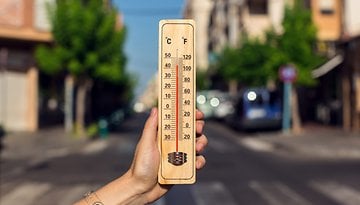 A hand holding a wooden thermometer showing high temperatures in Celsius and Fahrenheit on a sunny street.