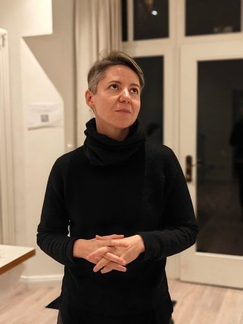 A person in a black outfit stands with hands clasped, looking thoughtfully in a well-lit room.