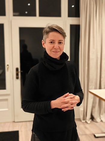 A woman with short hair in a black outfit stands with hands clasped, smiling in a well-lit room.