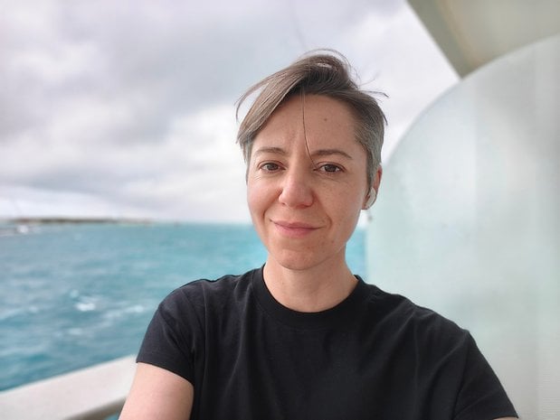 A person with short gray hair smiling on a boat with water and cloudy sky in the background.