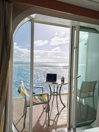 A balcony with a table and chairs overlooking the sea, with a laptop and a drink on the table.