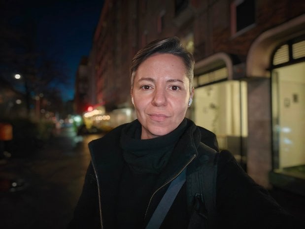 A person with short hair wearing a black jacket stands on a street at night.