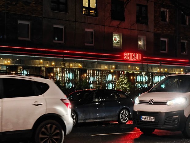 Uma cena movimentada da rua à noite com carros e um restaurante iluminado por luzes vermelhas.