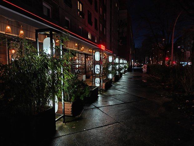 Vista noturna de um restaurante com lanternas brilhantes e plantas ao longo da calçada.