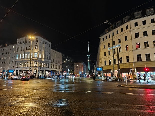 Eine belebte Straße bei Nacht, mit nassem Bürgersteig, beleuchteten Gebäuden und dem Fernsehturm.