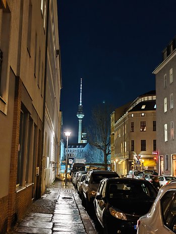 Vista noturna de uma rua com carros estacionados e uma torre ao fundo.