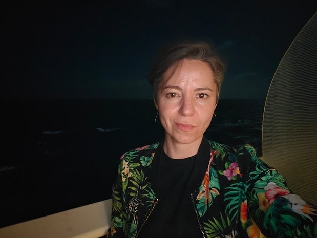 A woman with short hair wearing a floral jacket poses for a selfie at night near the ocean.