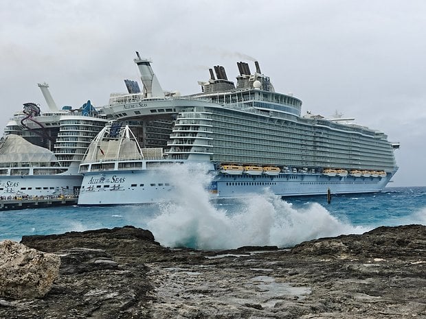 A large cruise ship, Allure of the Seas, docked near rocky shore with waves crashing in the foreground.