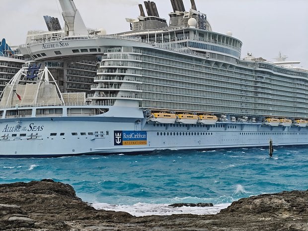 A large cruise ship, Allure of the Seas, docked near rocky shores with blue waters in the foreground.