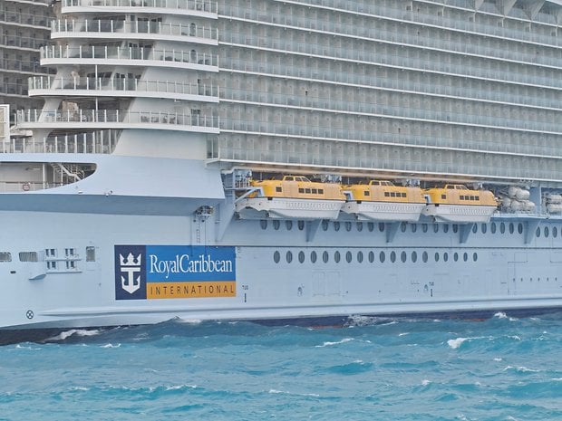 A large cruise ship with lifeboats, featuring the Royal Caribbean logo, docked in turquoise waters.