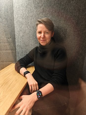 A person sitting at a wooden table, wearing a smartwatch and looking at the camera in a gray-walled room.