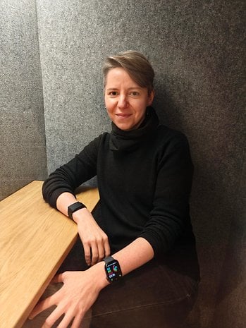 A woman with short hair sits at a table, wearing a black turtleneck and smartwatch.