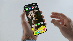Un iPhone vu de face, tenu dans une main gauche, avec son écran allumé affichant l'écran d'accueil d'iOS 18.