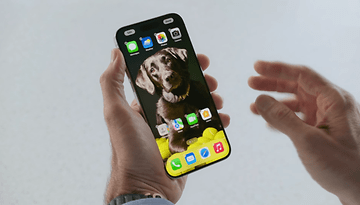 Un iPhone vu de face, tenu dans une main gauche, avec son écran allumé affichant l'écran d'accueil d'iOS 18.