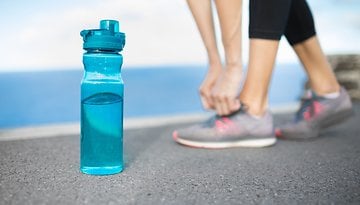 A blue water bottle sits on the ground, with a person tying their shoelaces in the background.
