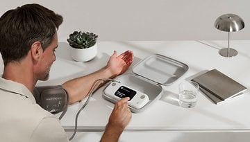 A man is using a BPM Vision device to measure his blood pressure at a desk.