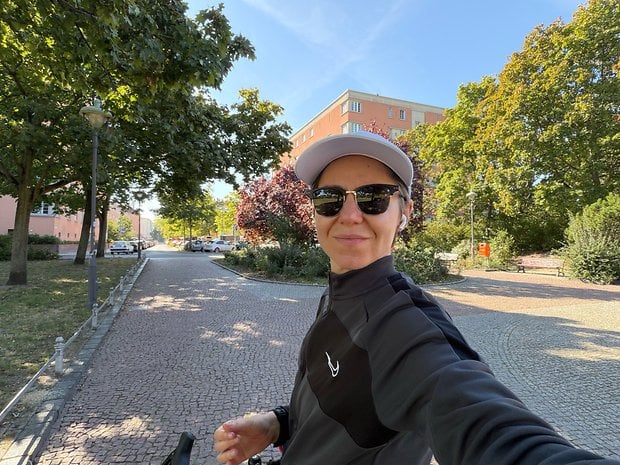 A person in a black jacket and sunglasses smiles for a selfie in a park with trees and buildings in the background.