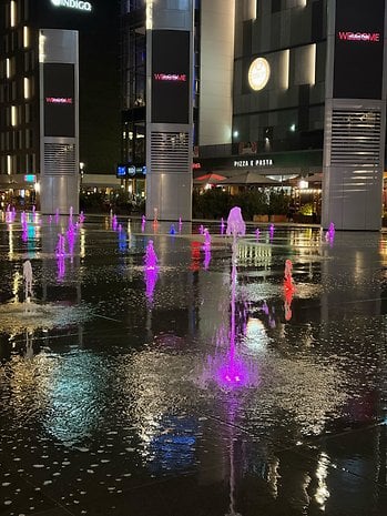 Fountains with colored lights in a plaza at night, featuring a 'WELCOME' sign.