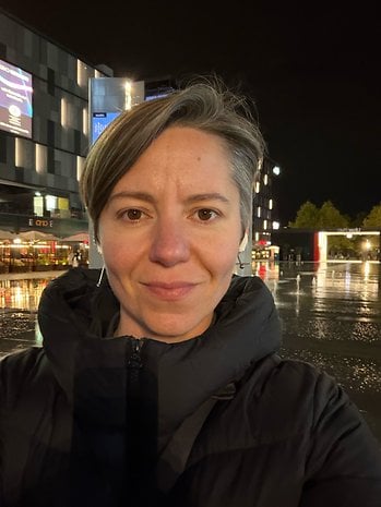 A person with short hair wearing a black jacket standing outdoors at night near illuminated buildings.