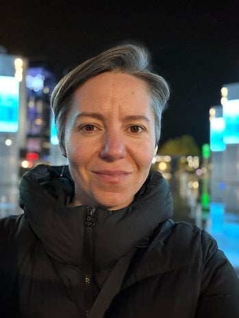 A woman with short gray hair, wearing a black puffer jacket, smiling at night with illuminated buildings in the background.