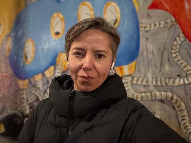 A person with short hair wearing a black jacket stands in front of a colorful mural.