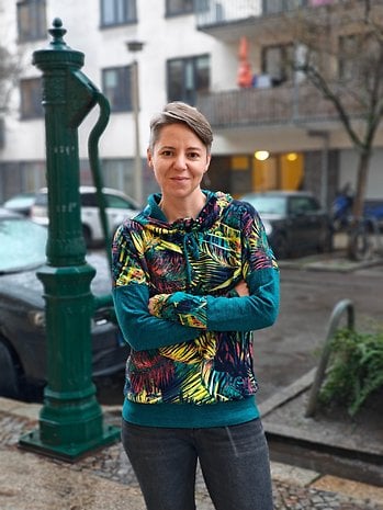 A person stands in front of a green water pump, wearing a colorful hoodie in a rainy urban setting.