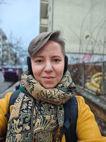 A person with short gray hair wearing a patterned scarf and backpack, standing outdoors on a cloudy day.