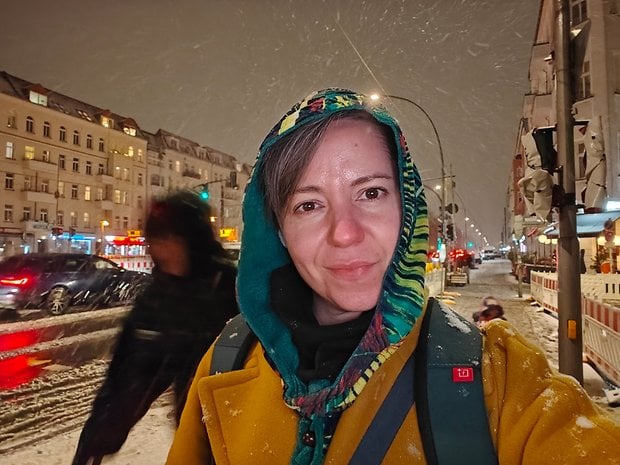 A person with a colorful hooded jacket stands in the snow at night, with city lights in the background.