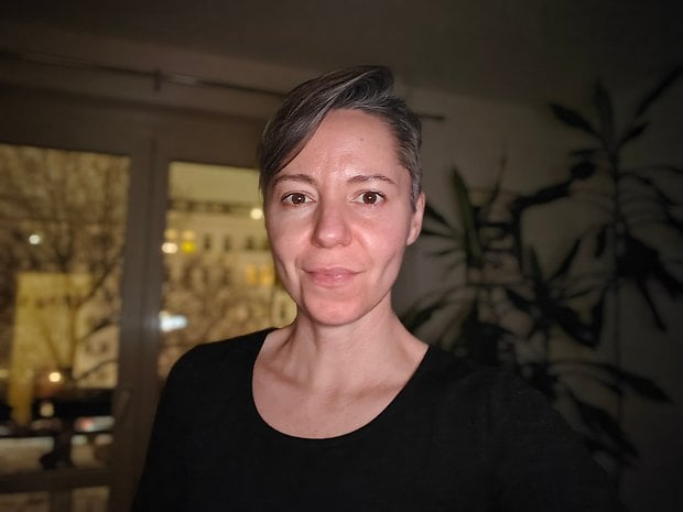 A woman with short hair stands indoors, smiling, with a nighttime city view behind her.