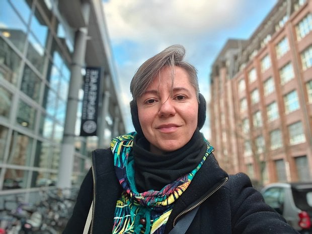 A woman with short hair wearing a colorful scarf and headphones, smiling outdoors near a building with bicycles.