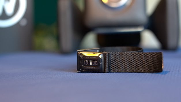 A close-up of a Whoop MG sensor on a black strap, placed on a textured surface.