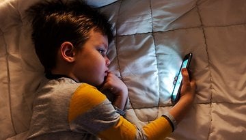 Uma criança deitada em uma cama, olhando para a tela de um smartphone na luz fraca.