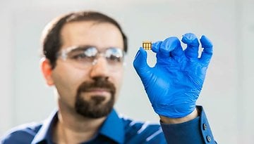 A researcher in a blue shirt and gloves holds a small solar cell designed for indoor light optimization.