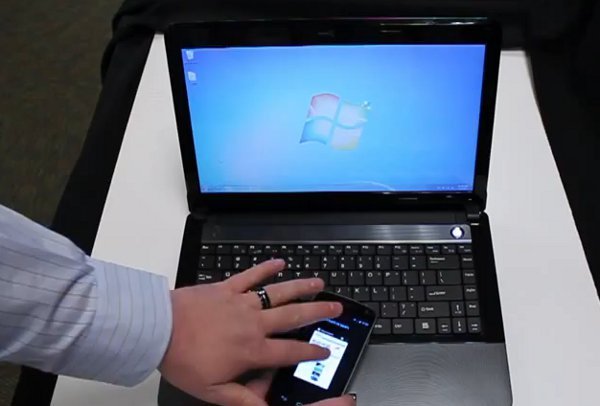 Une main posée sur un smartphone, près d'un ordinateur portable affichant le bureau Windows.