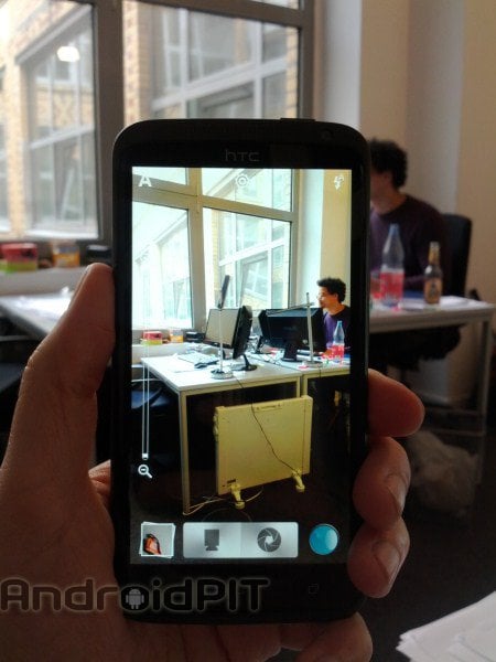 Un téléphone portable affichant une photo d'un bureau avec deux ordinateurs, un homme assis et une fenêtre.