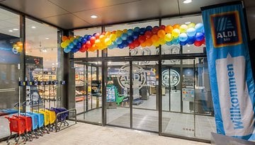 Eingang eines ALDI-Marktes, dekoriert mit bunten Ballons und einem Banner mit der Aufschrift 'Willkommen'.