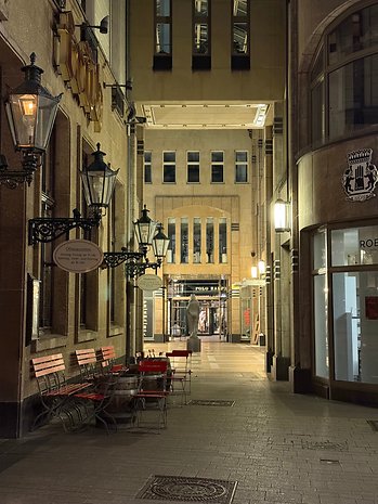 Eine schummrige Gasse mit roten Stühlen und Laternen, die zu einem Ladeneingang führt.