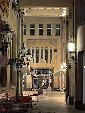 Eine nächtliche Gasse mit Geschäften, Straßenlaternen und einer Statue, die zu einem Polo Ralph Lauren Geschäft führt.