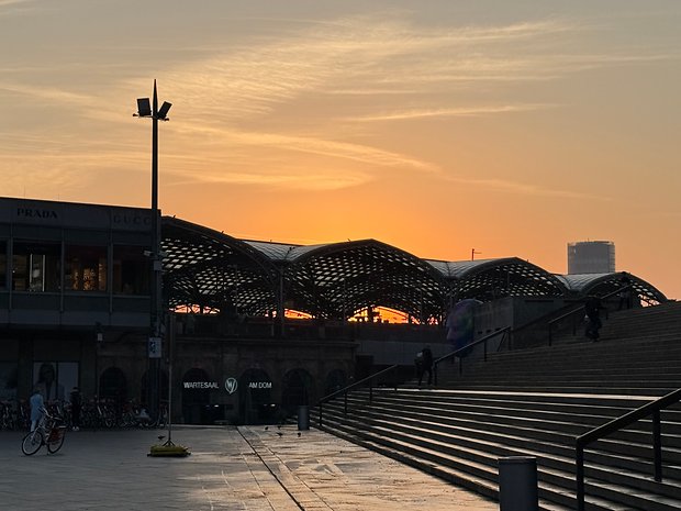 Pôr do sol atrás de uma arquitetura moderna com lojas, escadas e silhuetas de pessoas.