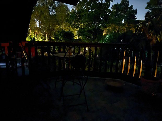 Uma varanda mal iluminada com uma mesa e uma cadeira com vista para um jardim à noite.