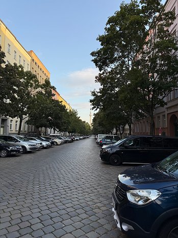 A cobbled road lined with trees and parked cars leading to a distant tower under a blue sky.