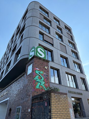 Um edifício moderno com fachada curva e um sinal verde 'S', apresentando grafite na parede de tijolos.