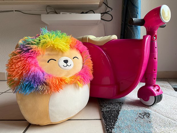 A colorful plush lion next to a bright pink scooter on a tiled floor.