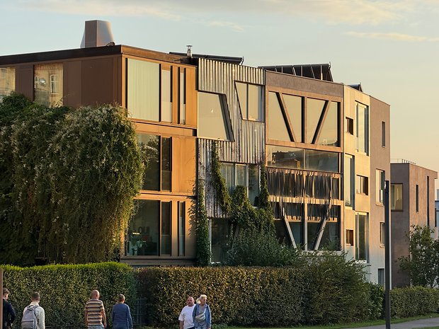 Modern buildings with large windows and green spaces, people walking around during sunset.