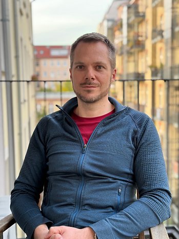 Ein Mann sitzt auf einem Balkon, trägt eine blaue Jacke, mit einer Stadtansicht im Hintergrund.