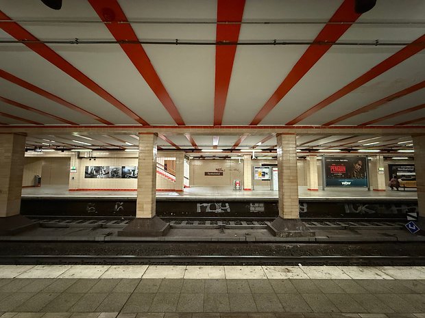 Uma estação de metrô com tetos listrados em vermelho e branco, plataformas vazias e anúncios nas paredes.