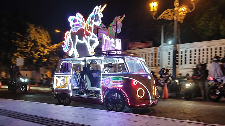 A brightly lit vehicle with a unicorn on top driving at night, passengers visible inside.