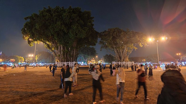 Night scene of a park with large trees and people walking, illuminated by streetlights and festive lights.